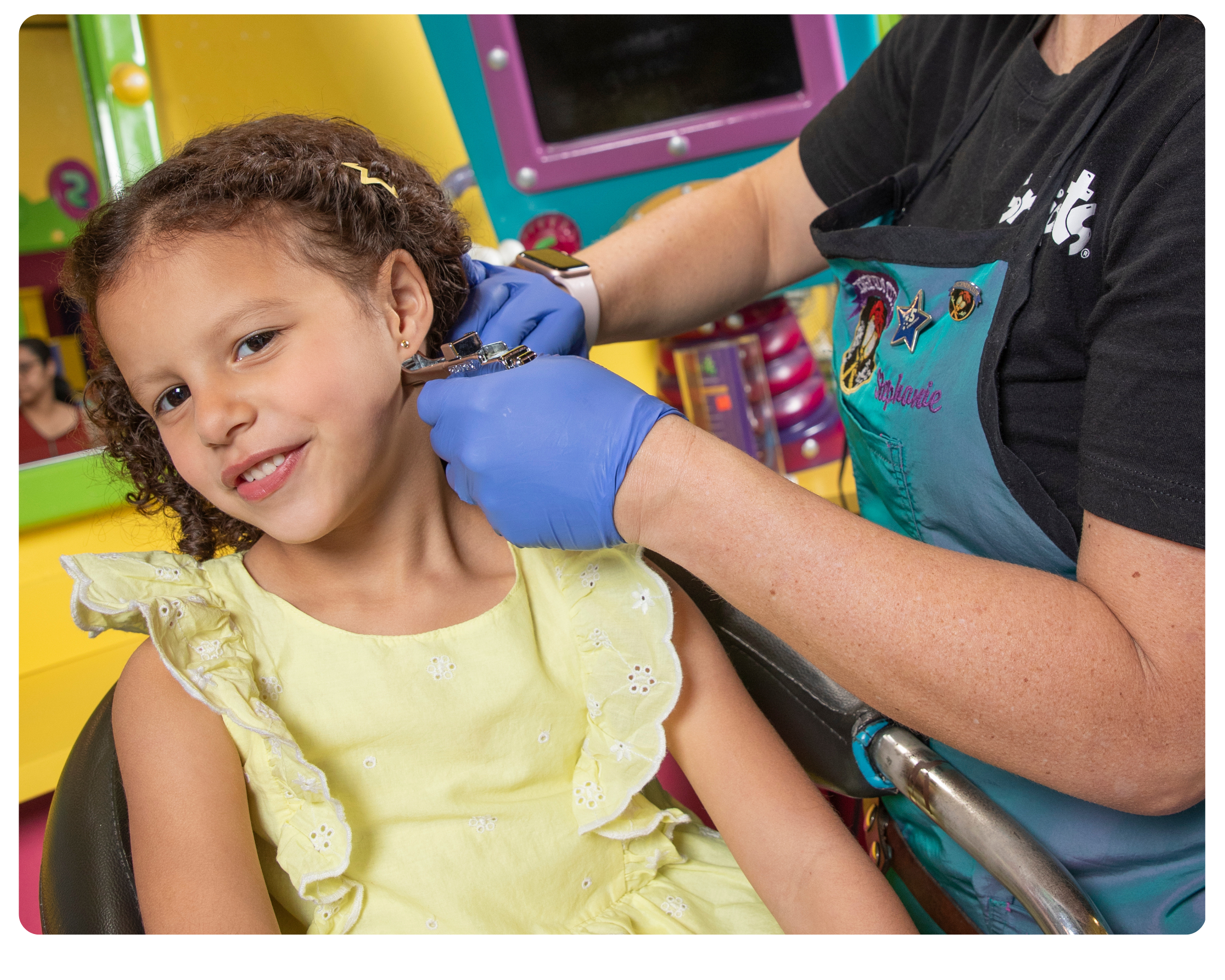 A girl in a yellow dress sits calmy in a salon chair whilst her ears are getting pierced.