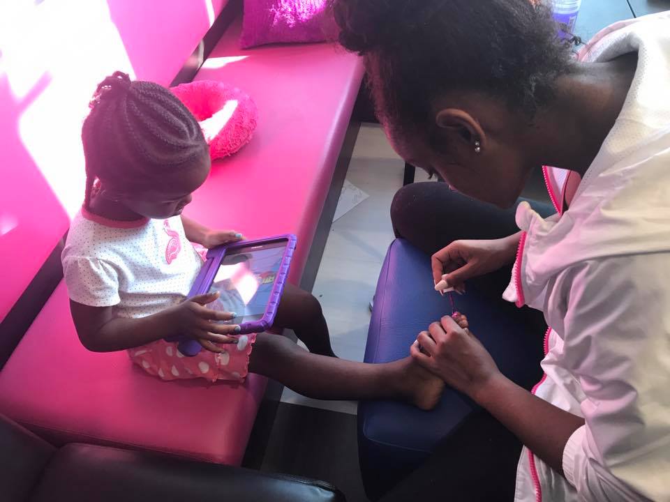 A woman painting the toes of a girl sitting on a pink bench with a purple tablet in her lap.