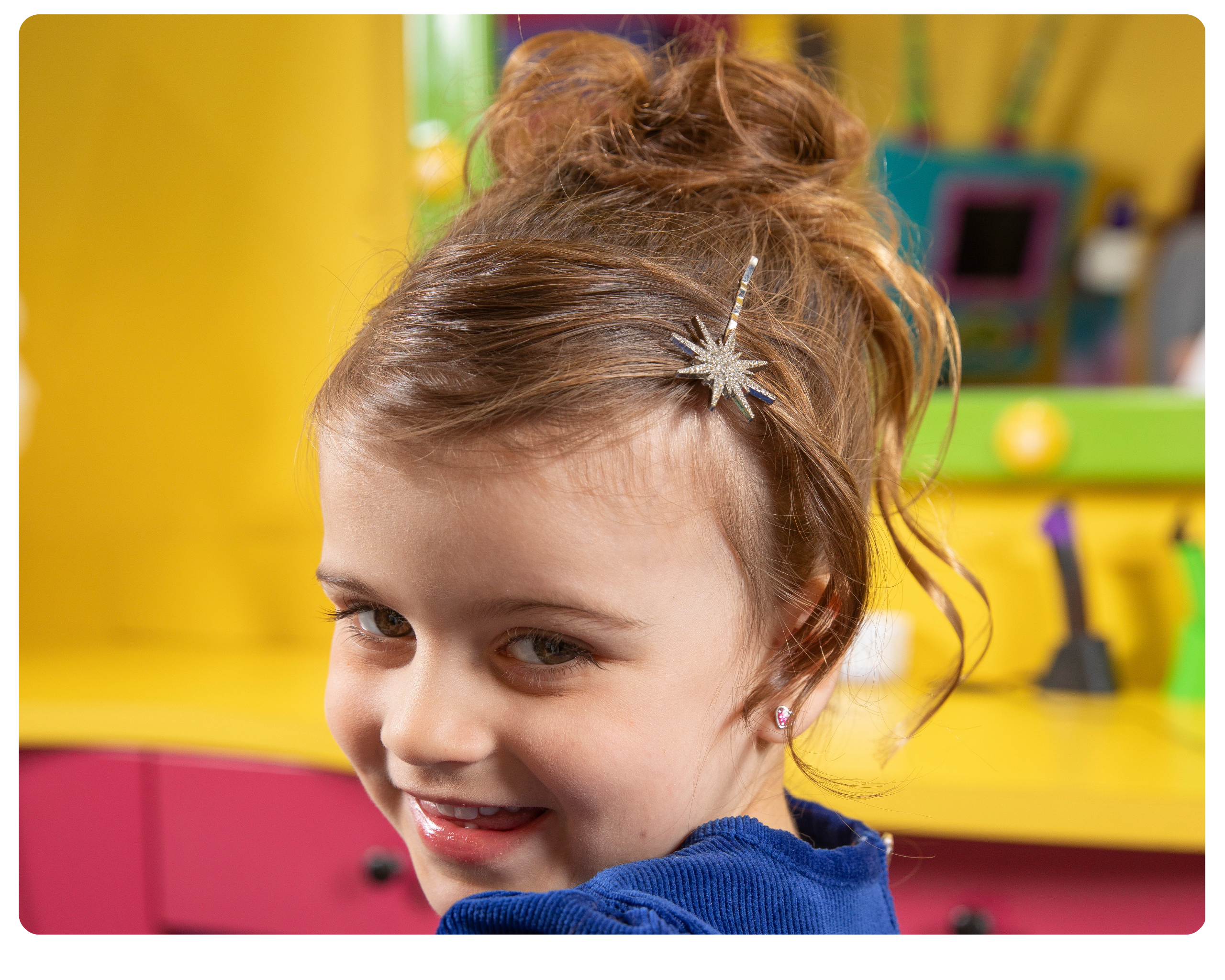 a girl with her hair up and a diamond starburst hair clip in her bangs, smiles at the camera against a yellow and pink back ground.