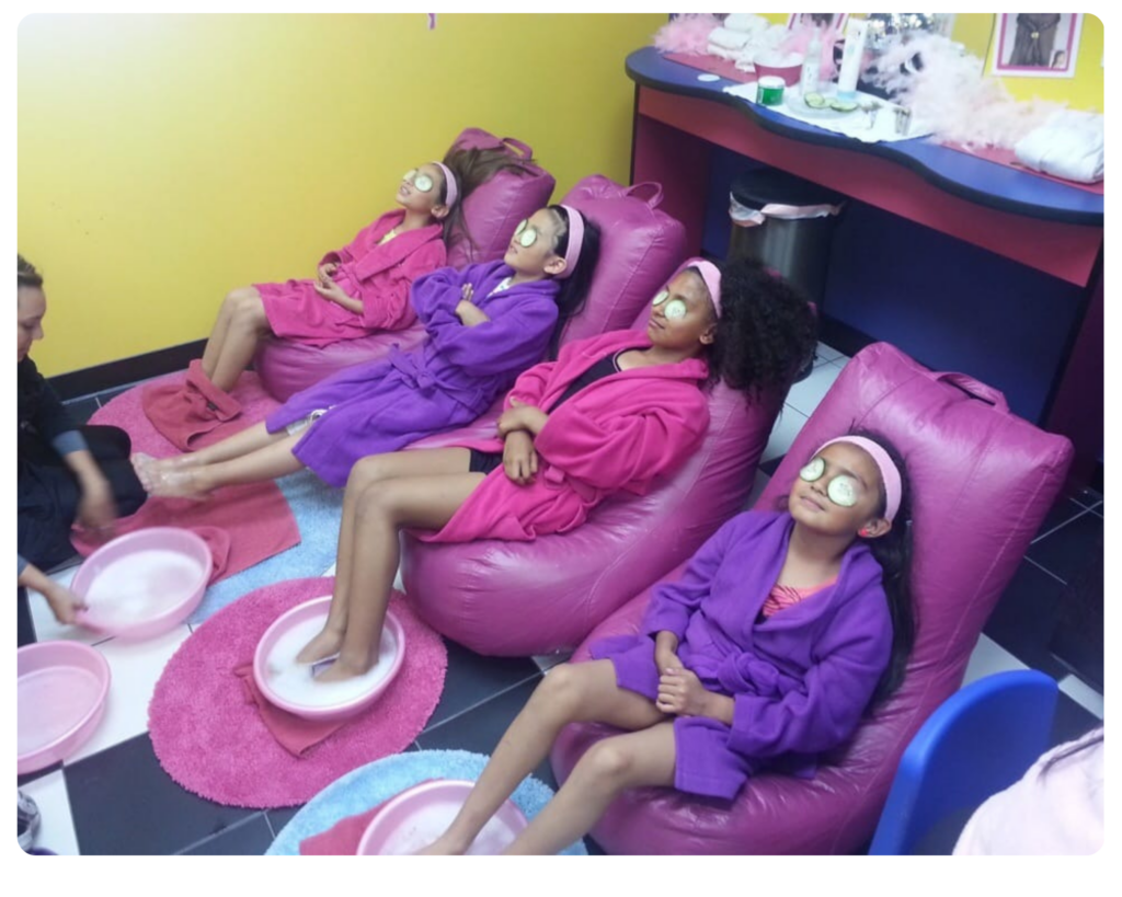 Four girls in pink beanbag chairs wearing spa robes and soaking their feet in pedicure bowls. Cucumbers on their eyes and spa headbands in their hair.