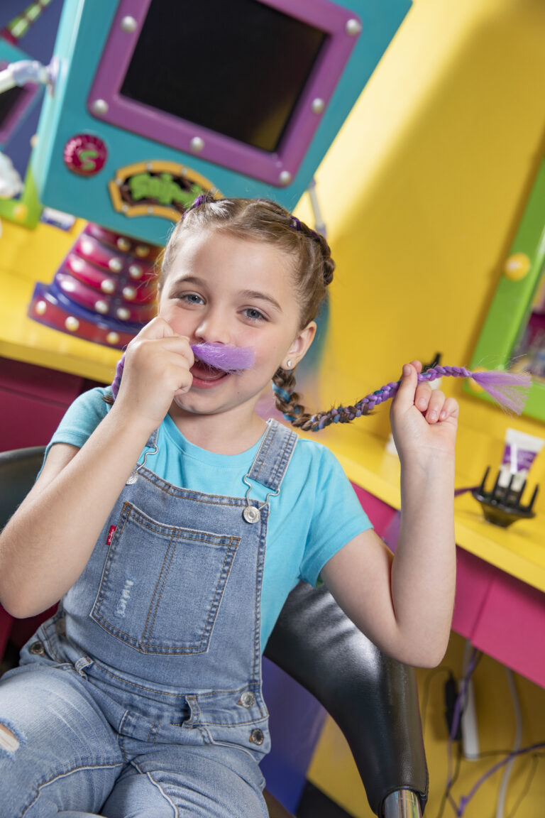 a girl in denim overalls holding onto her braids whilst sitting in a salon chair.