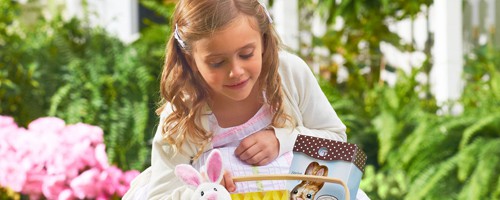 A girl with blonde hair looking in her Easter basket that has an Easter bunny toy and chocolate bunny.