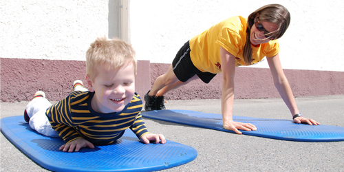 A young boy playing on a yoga mat next to his mom outside.