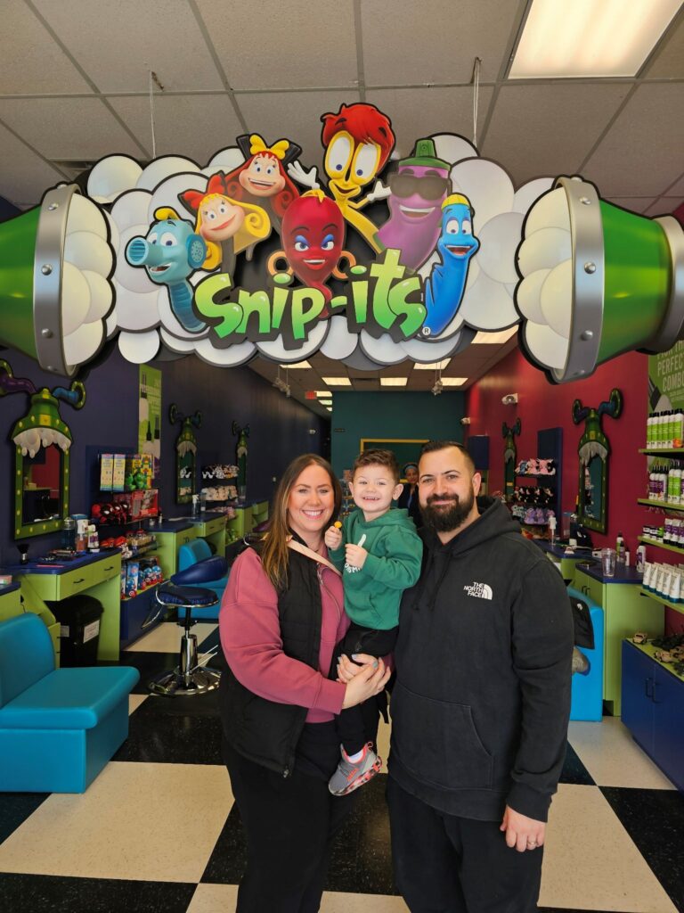 A smiling boy between his two parents at Snip-its Haircuts for Kids in West Caldwell, NJ. The salon's first customers standing underneath the Snip-its character arch.